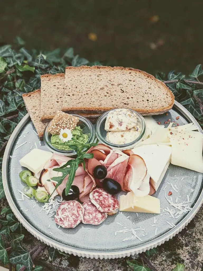 Brotzeitplatte mit Käse, Wurst, Brot und verschiedenen Beilagen auf einem runden Teller.