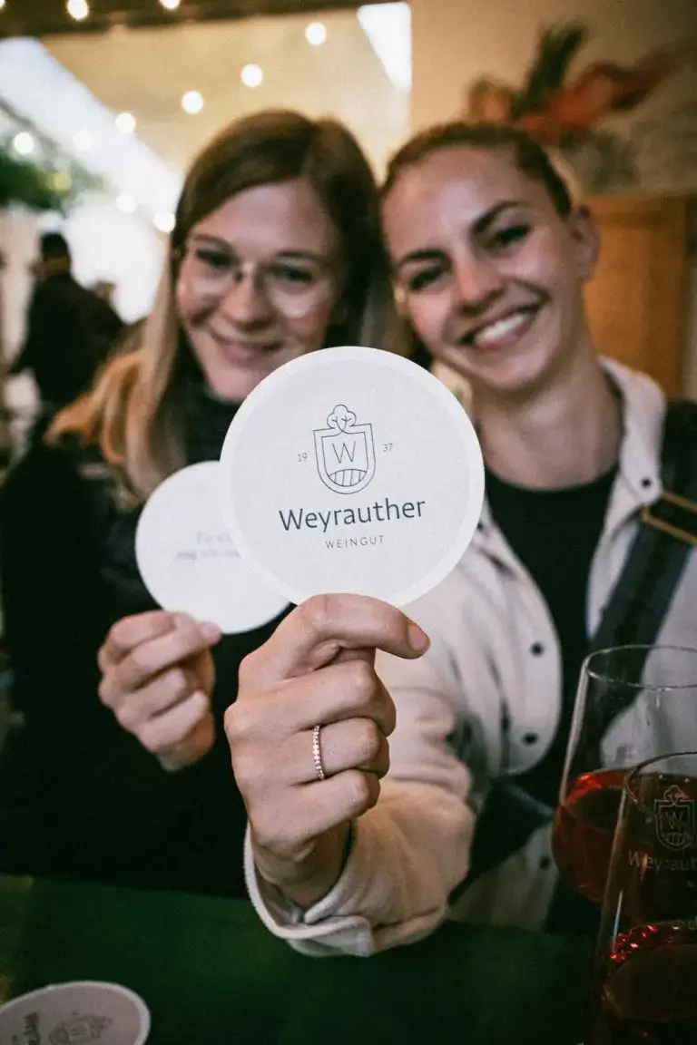 Zwei Personen halten runde Untersetzer mit dem Logo des Weinguts Weyrauther.